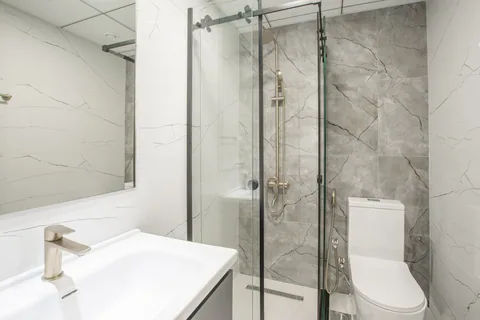 Baño de apartamento con lavabo, mampara de ducha de cristal e inodoro con cisterna, en tonos blancos y grises