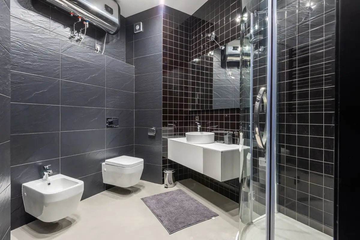 Termo eléctrico colgado en lo alto de la pared de un baño alicatado en gris oscuro, con sus tomas de agua